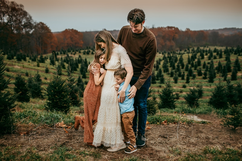 Family Photographer, mom, dad, daughter, and son huddle close before a a tree farm