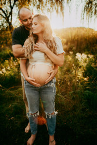 Maternity Photographer, Man embraces pregnant woman from behind, she leans into him, they are beneath a tree